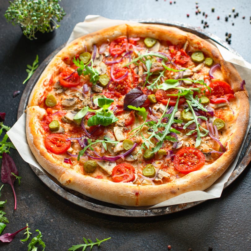 A freshly baked pizza topped with tomatoes, mushrooms, red onion, pickles, and arugula.