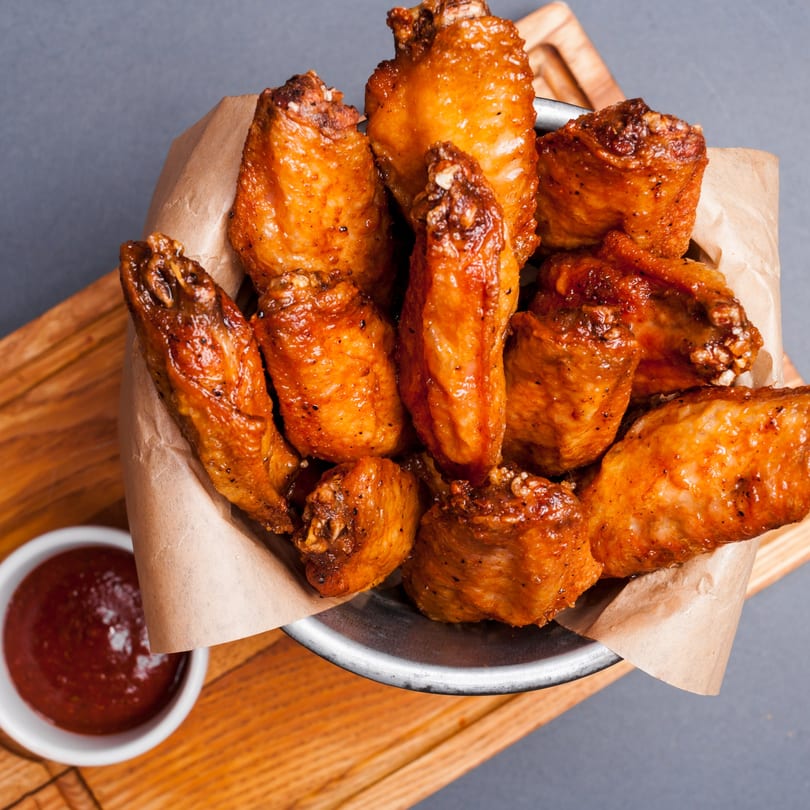 Crispy, golden-brown chicken wings in a metal bowl lined with parchment paper, with a side of dipping sauce.